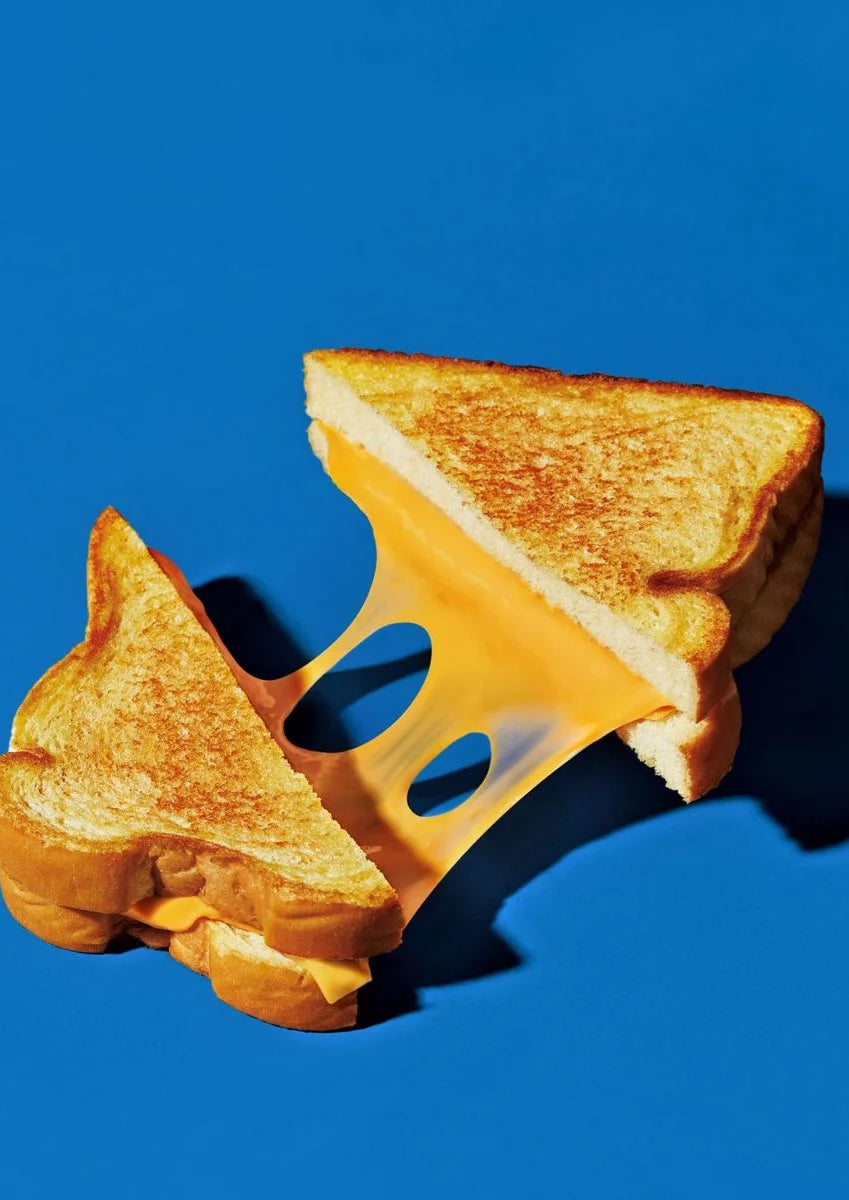 Contraste saisissant entre des pains dorés chauds et une toile bleue froide capture un grilled cheese fendu avec du fromage étiré, évoquant des textures riches et une chaleur appétissante.. blue, photographic, still life, warm, white, yellow