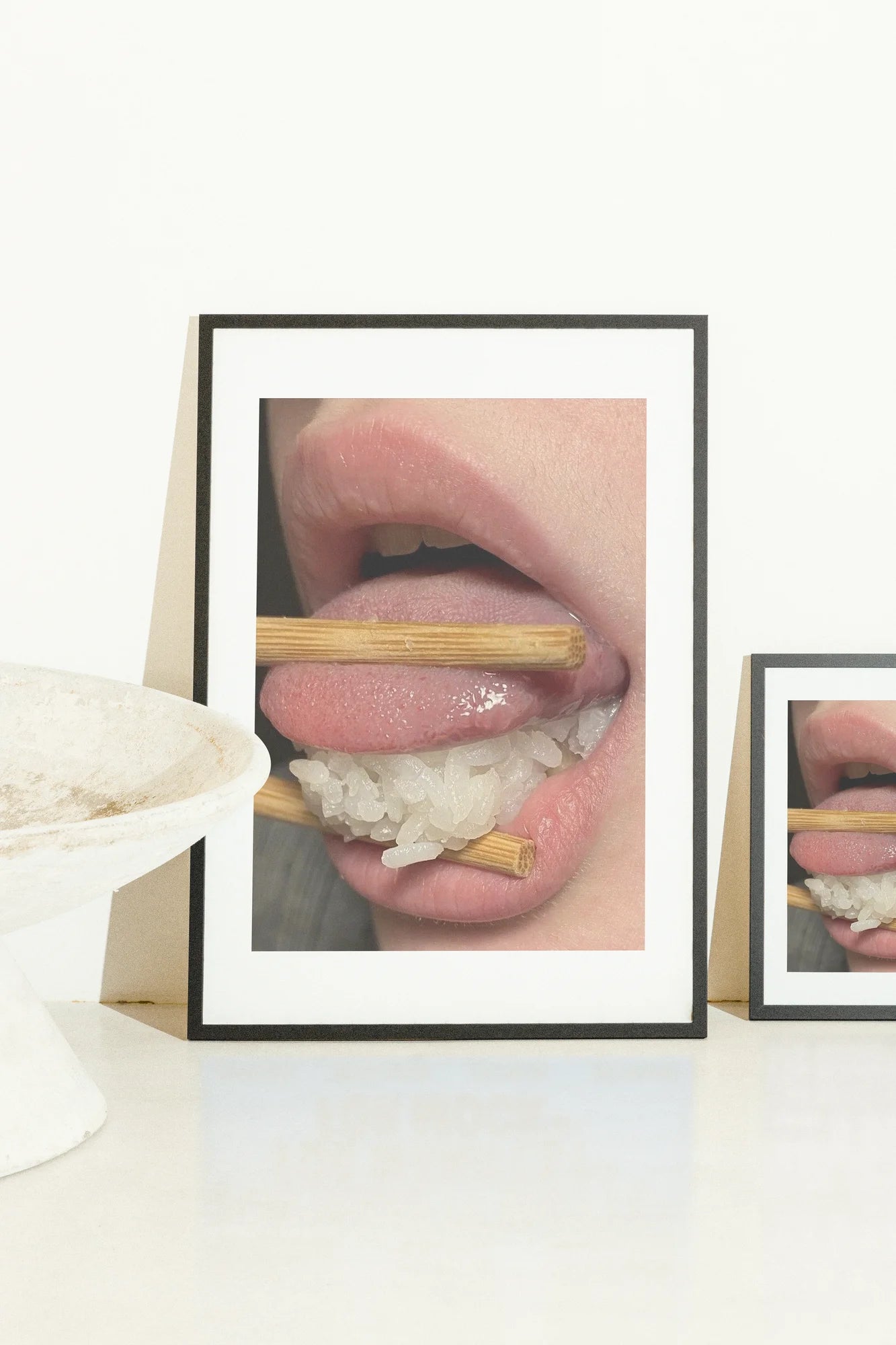 Photographie de la bouche d'une personne tenant des baguettes avec du riz ; les tons de peau naturels, les baguettes marron chaudes contrastent avec le riz blanc, créant un gros plan intime.. black, brown, photographic, portrait, still life, white